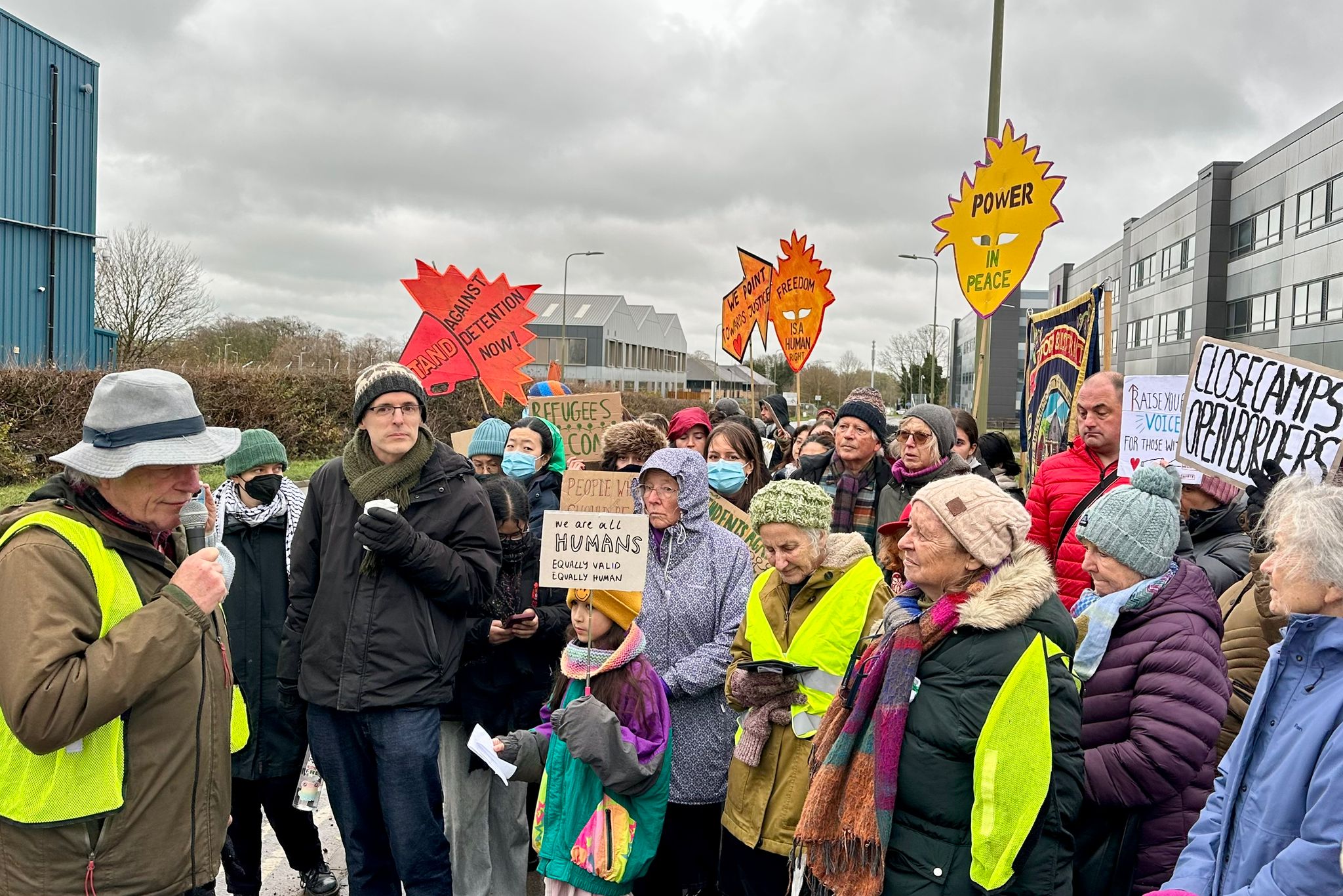 14 Roadside view with a sign pointing to “Ambulance Station” and “Campsfield House” on the left, while a large group of protesters with placards gathers on the pavement in the distance outside the Campsfield site under an overcast sky.