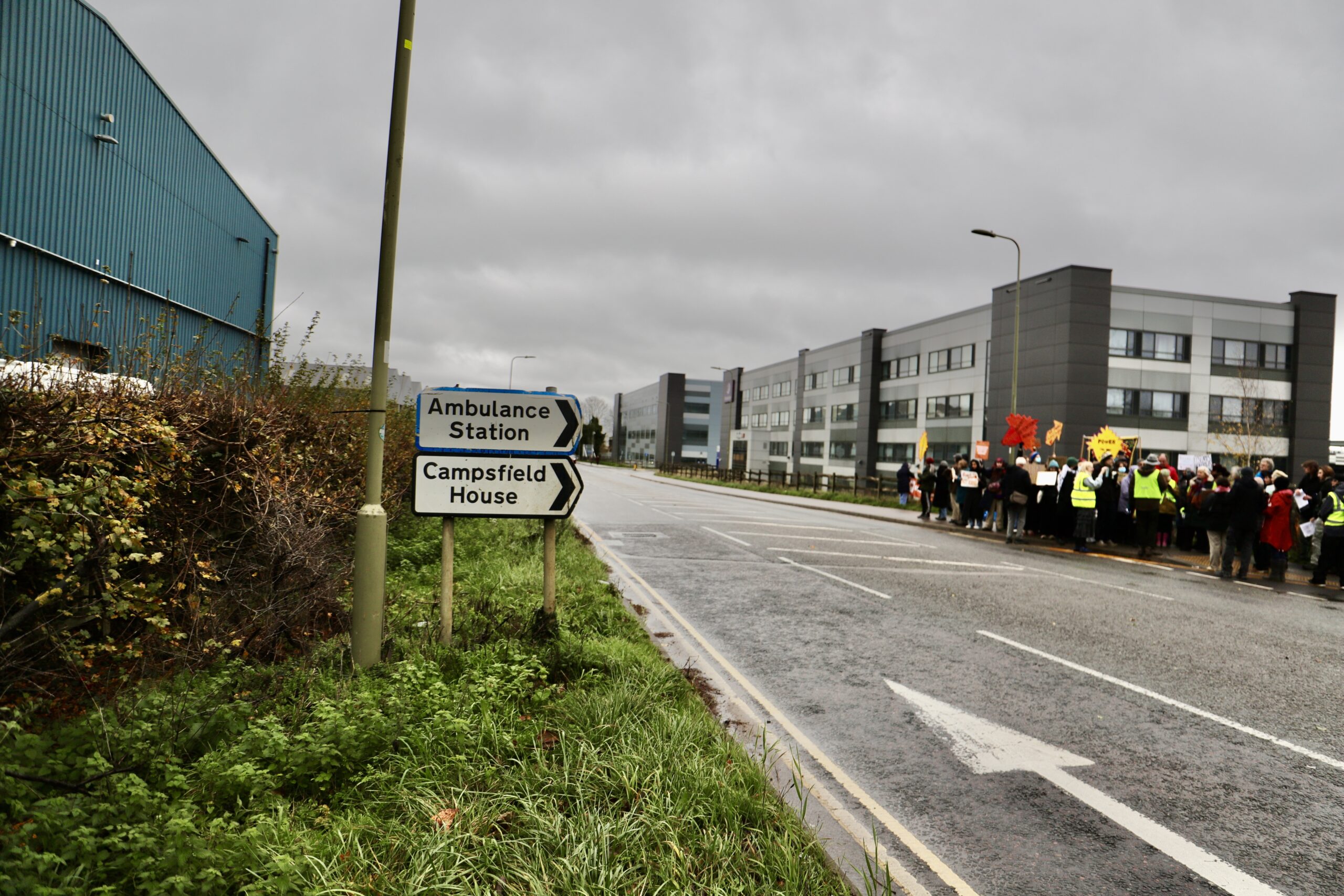 19 Roadside view with a sign pointing to “Ambulance Station” and “Campsfield House” on the left, while a large group of protesters with placards gathers on the pavement in the distance outside the Campsfield site under an overcast sky.