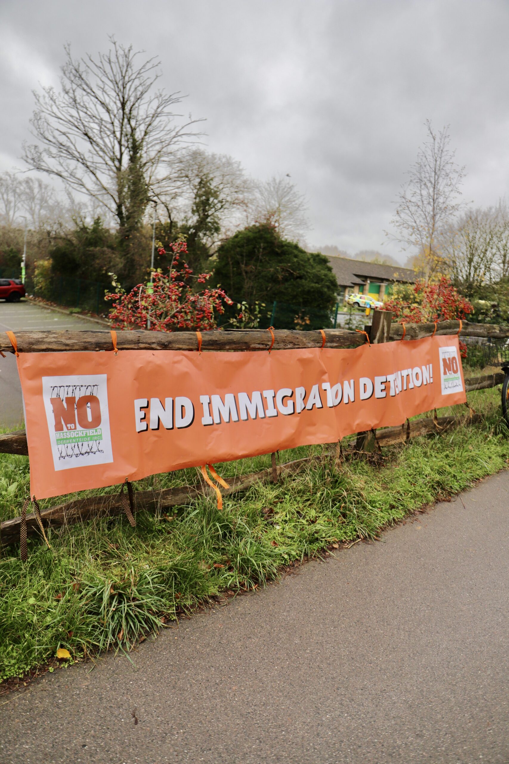 An orange banner reading “END IMMIGRATION DETENTION” with “NO” symbols at each end is tied to a wooden fence along a roadside verge, with hedges, trees and a cloudy sky in the background