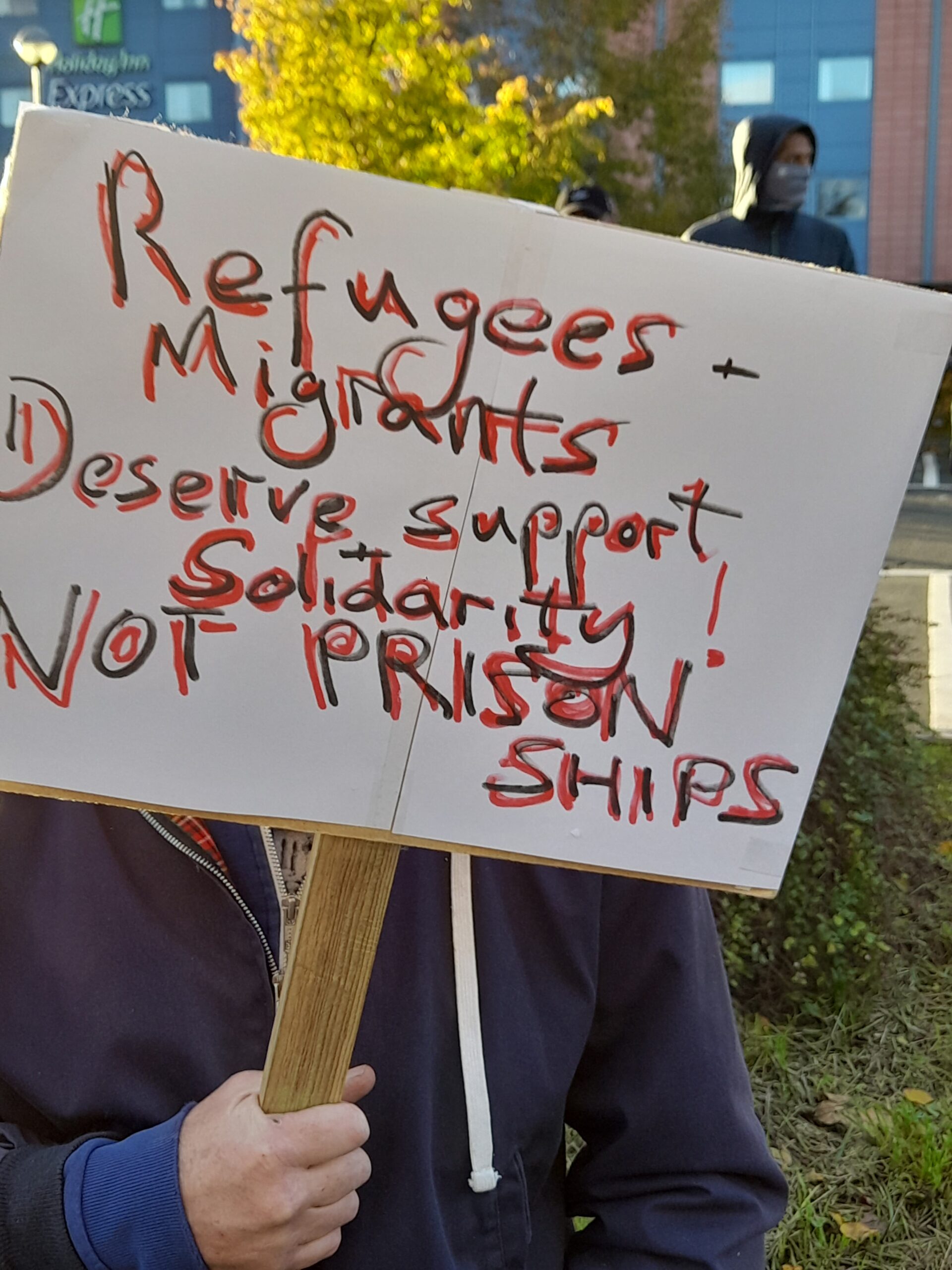Placard reads "Refugees + migrants deserve support + solidarity! NOT PRISON SHIPS"