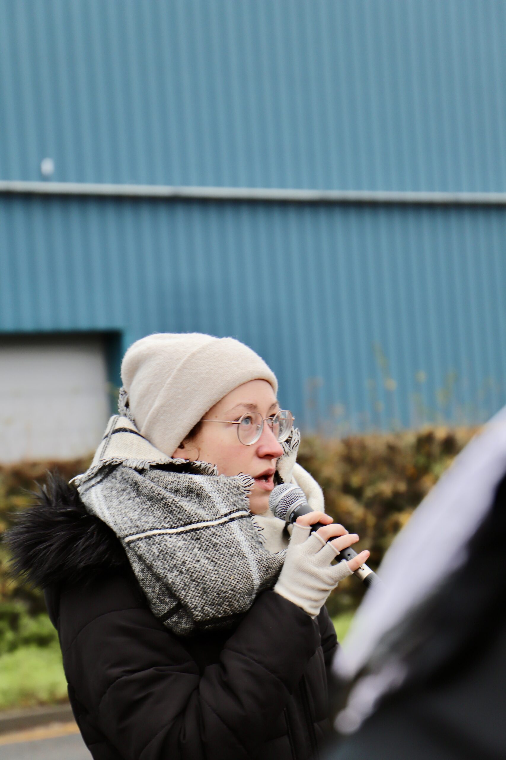 40 A speaker wearing a beige beanie, glasses and a thick scarf holds a microphone and addresses the crowd outside Campsfield, with blue industrial buildings and winter hedges in the background.