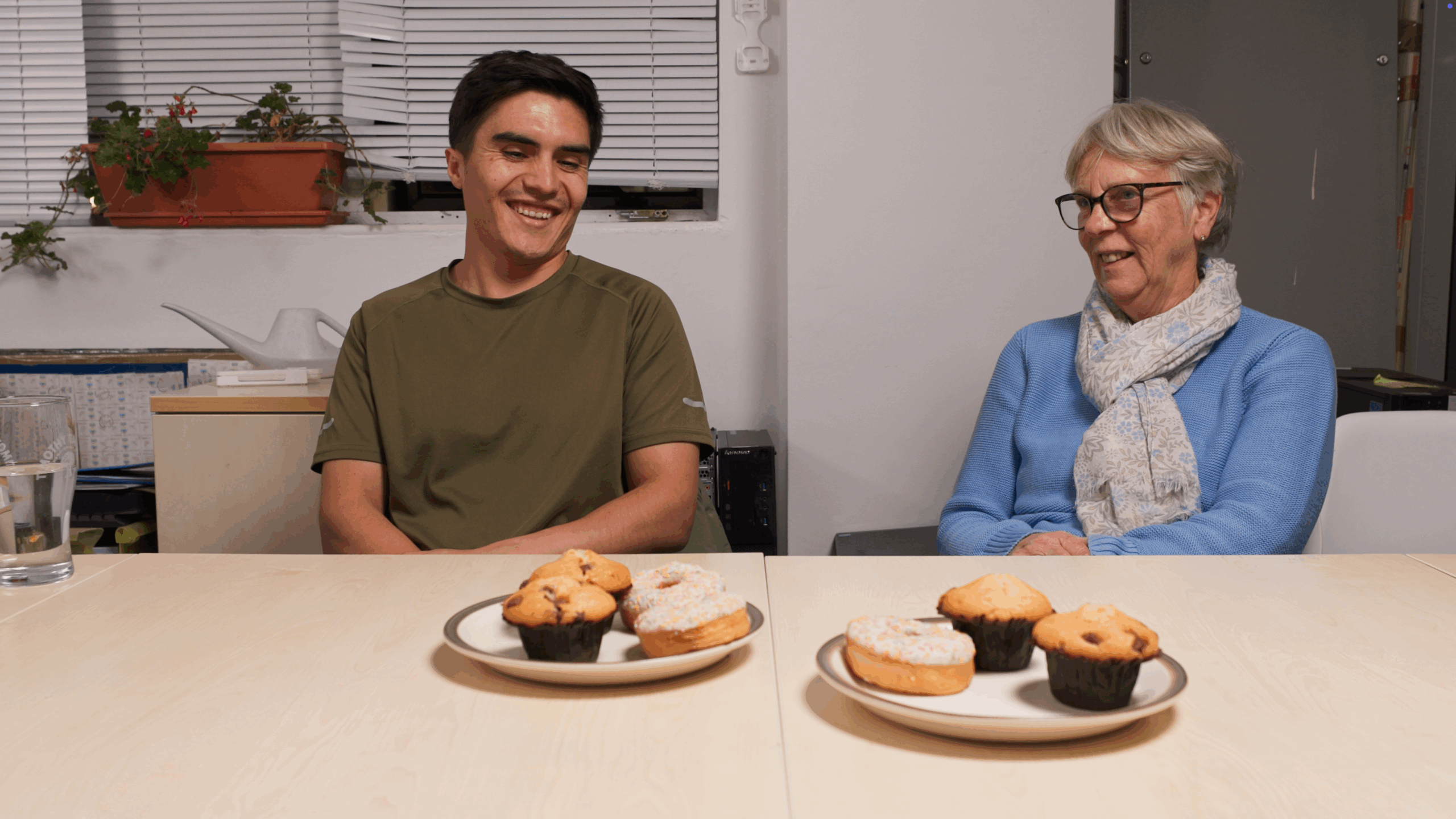 A young man and an older woman sit at a table laughing together, with plates of muffins and doughnuts in front of them. A plant and watering can are on the windowsill behind.