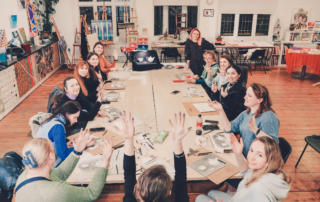 A group of Ukrainian women gathered around a table at an art workshop, listening to a teacher.